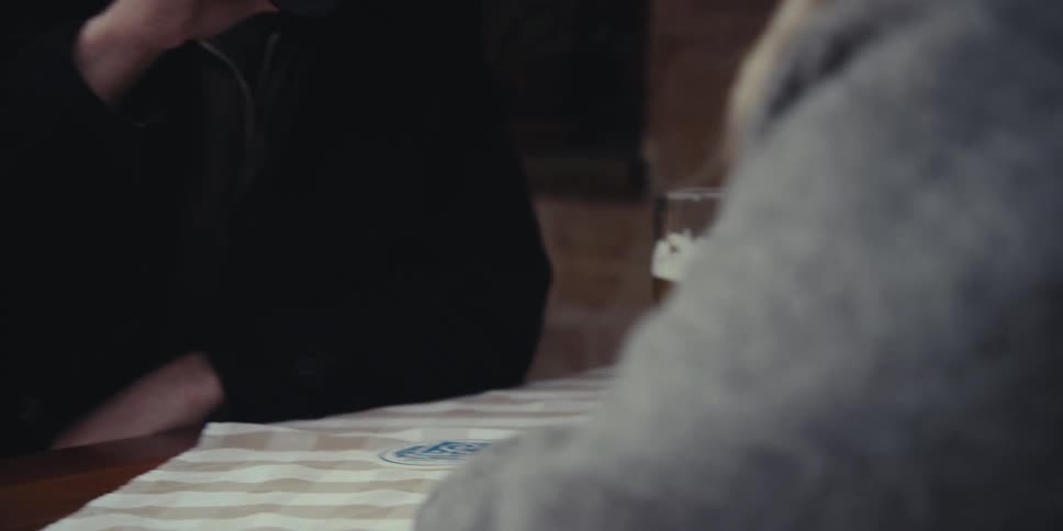 A man and a woman share a brief intimate moment at an outdoor table as he drinks a dark beer and their hands touch