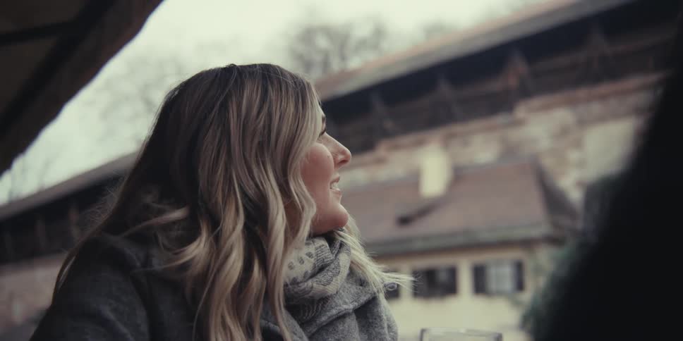 A smiling woman wearing a grey coat and scarf holds a glass of beer while talking with friends at an outdoor table in front of a stone building with an arched gateway