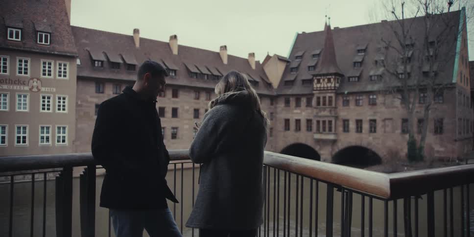 A couple stands on a balcony overlooking a river and historic european buildings after talking the woman pulls out her smartphone and the pair smiles for a selfie together