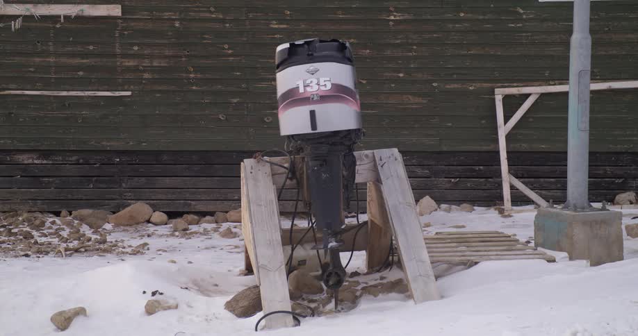 An old black and silver outboard motor with the number 135 sits mounted on a makeshift wooden stand in a snowy outdoor area next to a dark wooden building and various scattered debris