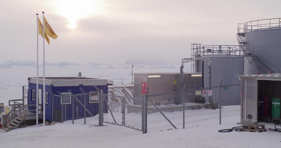 Flags and a blue and white building stand near a fenced lot containing multiple oil tanks on an overcast winter day
