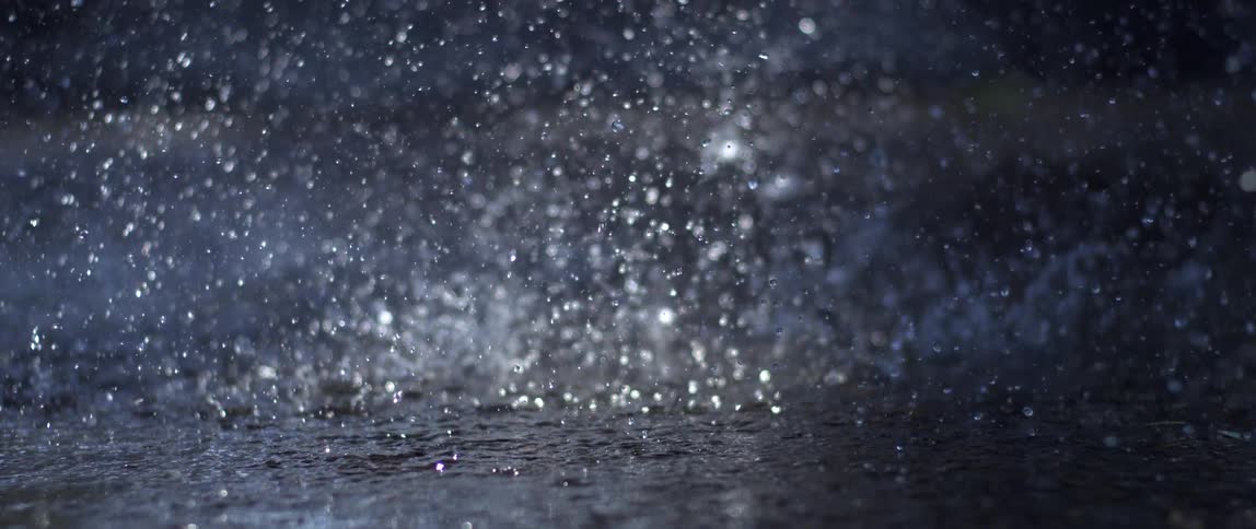 Heavy rain splashes onto a dark wet surface in slow motion creating intricate crowns and ripples the scene is dimly lit with a blue tint highlighting the shimmering droplets against a soft outoffocus background