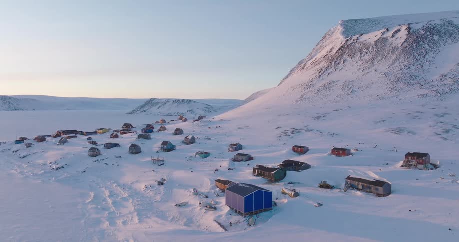 An aerial shot shows a cluster of colorful houses in a remote arctic village nestled at the base of a large snowy mountain during a clear winter day
