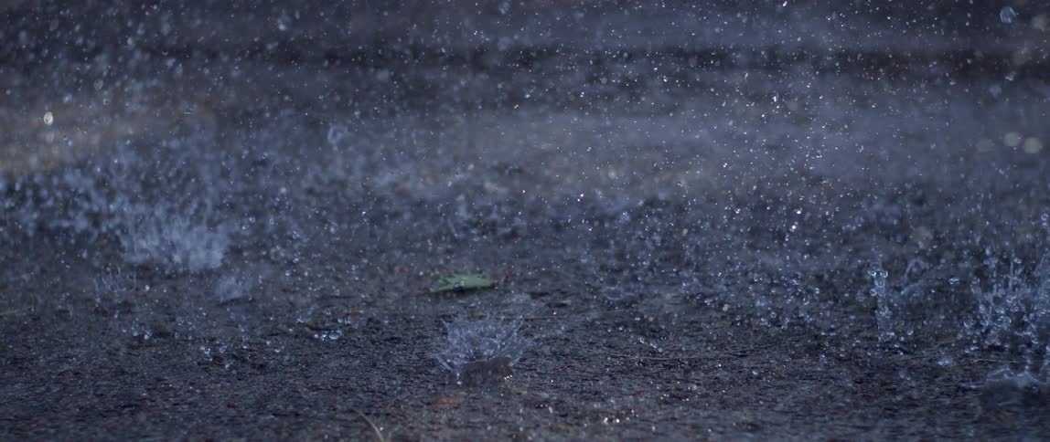 Heavy rain pummels the dark asphalt the droplets splashing vigorously upon impact and creating a mesmerizing rhythmic texture across the wet surface