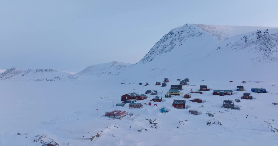 A small town sits on a snowy hillside at midday with scattered buildings along the slope and a snowcovered mountain rising in the background