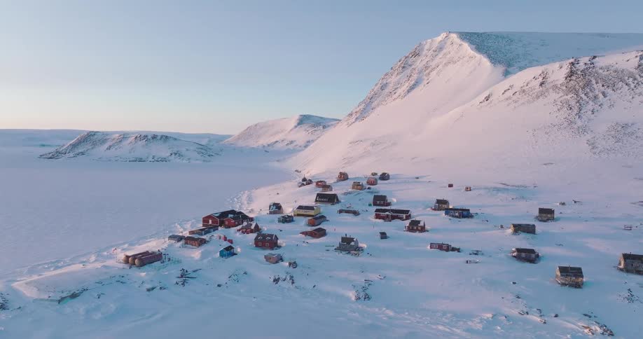 An aerial view lowers gracefully over a remote collection of colorful huts nestled on a snowy slope set against a majestic backdrop of icy mountains and soft sunset light