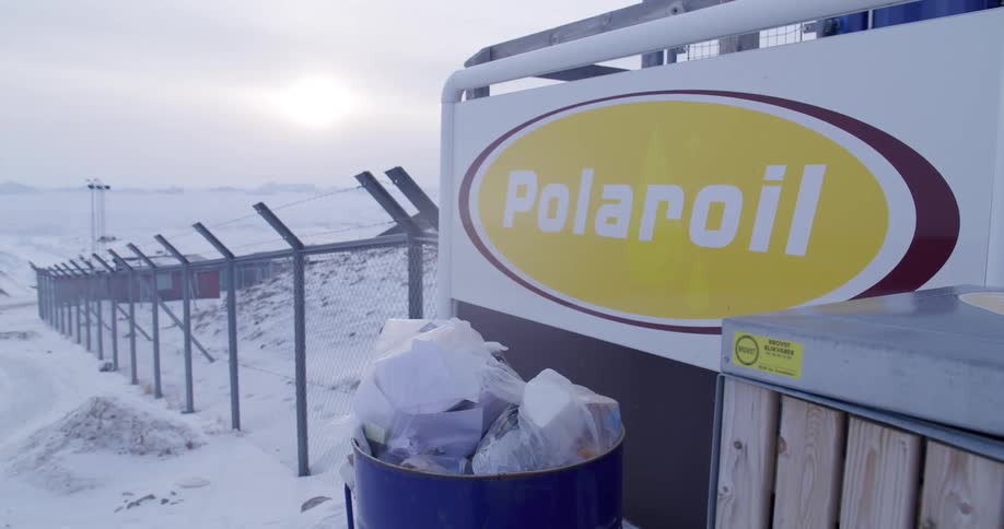 An overflowing trash bin sits in front of a polaroil sign at a fenced industrial site in a snowy arctic landscape buildings are visible behind a chainlink fence topped with barbed wire under a pale hazy sky