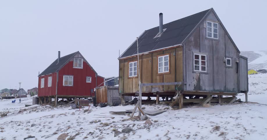 Two small weathered houses one red and one greyishbrown sit on stilts in a snowy desolate landscape under a grey sky