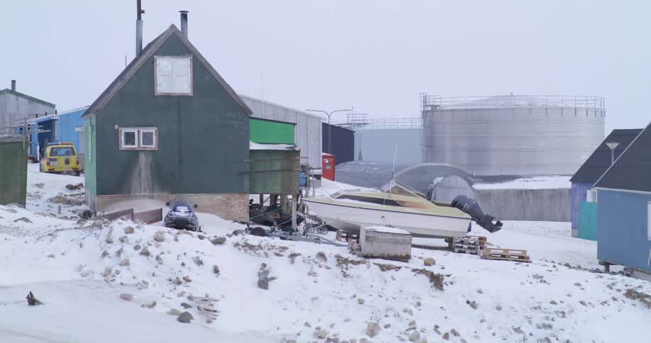 House in a polar settlement with a boat resting on snowcovered ground and industrial tanks in the background