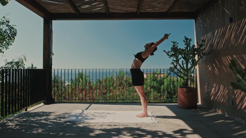 On a sundappled patio with a vast ocean view a woman gracefully flows through a sun salutation reaching her arms to the sky and arching back before folding forward toward her mat