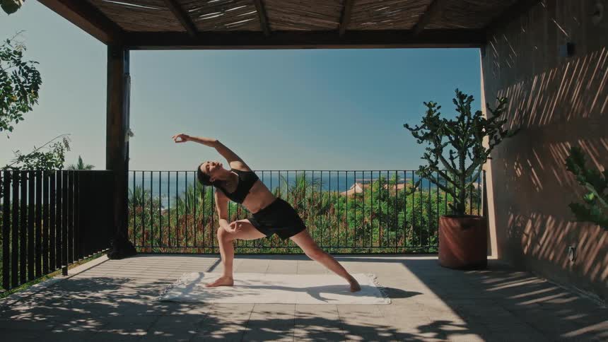A woman gracefully transitions from a strong warrior pose into a deep side angle stretch her movements fluid and controlled on a sunlit patio overlooking a lush coastal landscape