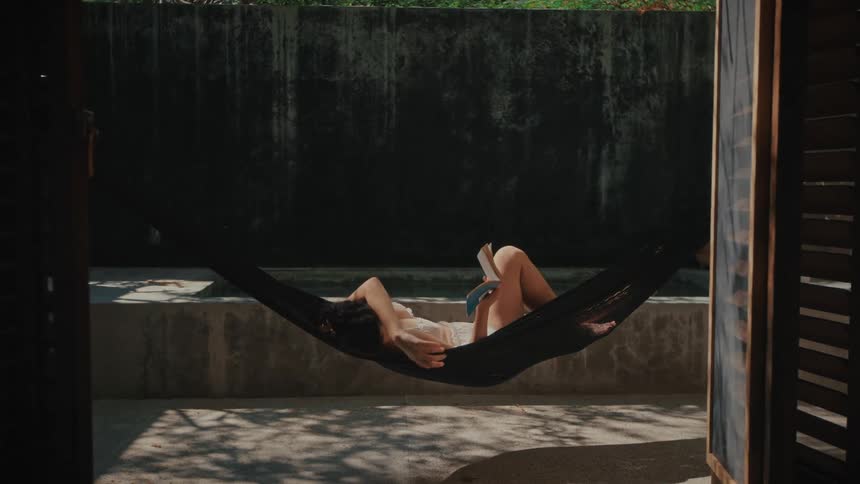 A young woman lies on a hammock on a sunny patio reading a book as the light falls directly across her