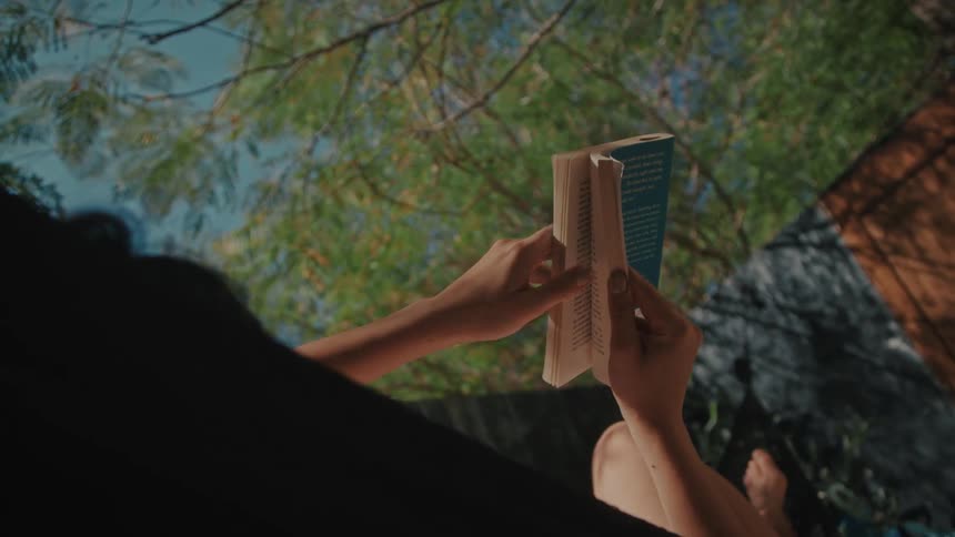 From a low angle a person relaxes outdoors under leafy green trees leisurely flipping through the pages of a book as sunlight filters gently through the branches above