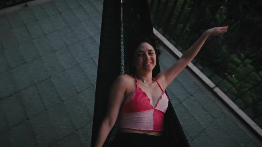 Young adult woman lying down on a hammock and pushing with her hand against a handrail in a balcony outdoors during dusk or dawn
