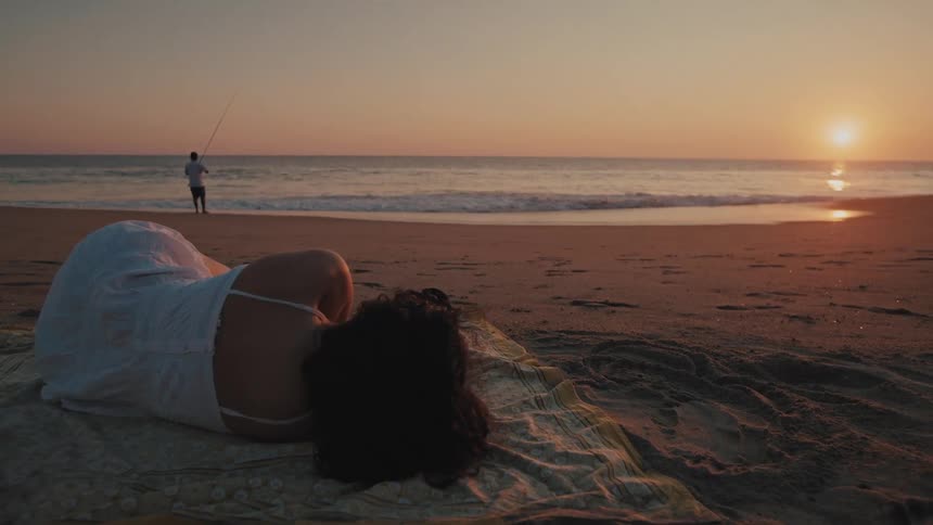 Woman lying on a towel on the beach at sunset with a person fishing in the background