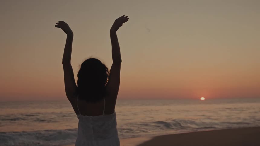 A young woman dances ballet on the beach at sunset her hands lifted gracefully as waves roll in and break along the shoreline