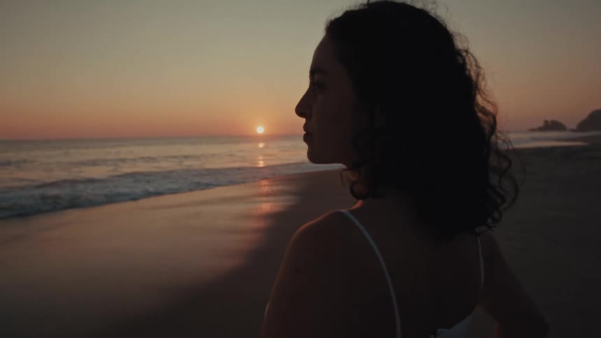 Young adult female wearing a white dress looking at the ocean on a beach outdoors during dusk or dawn