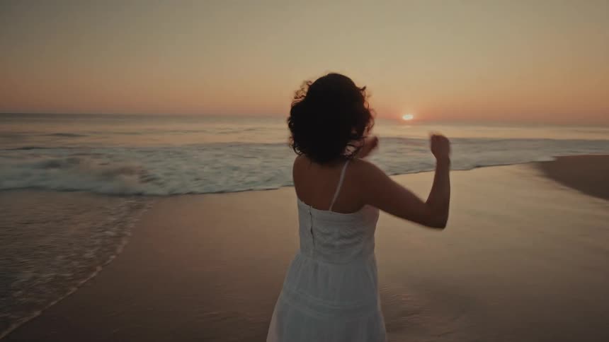 A woman in a white dress walks through the shallow surf and turns to look at the sunset over the ocean