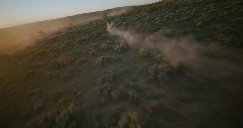 Two bikers on motorcycles riding on a dust road surrounded by trees and vegetation with hills on the far distance outdoors during dusk or dawn