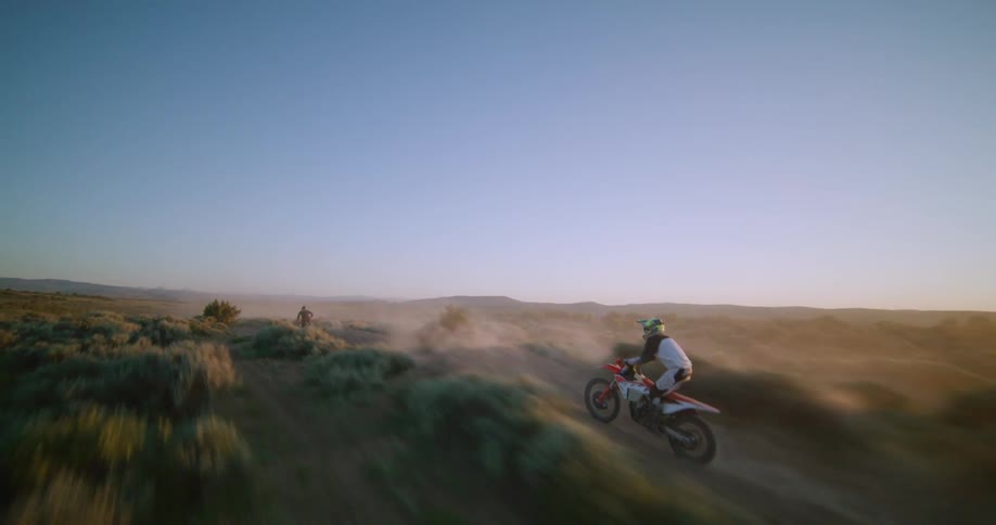 A dynamic aerial shot tracks motocross riders as they carve down a winding dirt hill launching over dramatic jumps and kicking up plumes of dust against the golden sunset