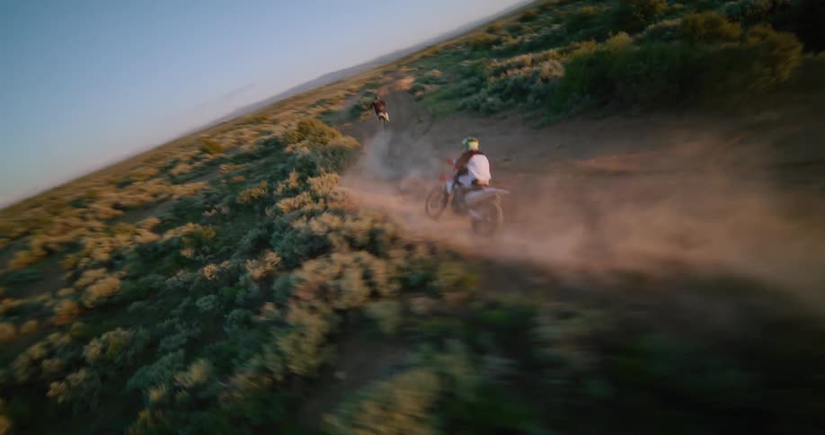 Two people on motorcycles ride across the rural terrain leaping off hills
