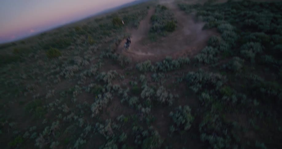 A motorcyclist speeds along a winding dirt trail through a vast desert landscape at twilight as a highspeed drone tracks their aggressive maneuvers sharp turns and jumps through the low light