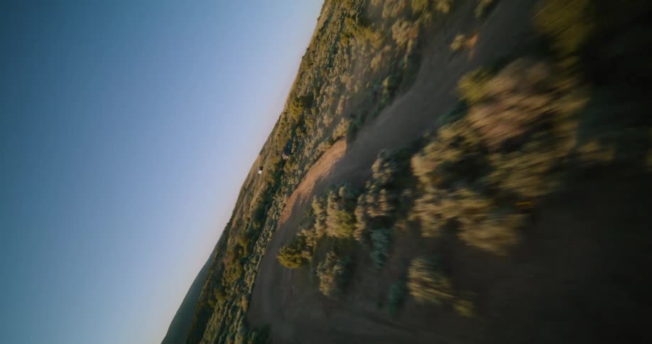 A motorcyclist rides across a dusty open desert landscape at bright sunset