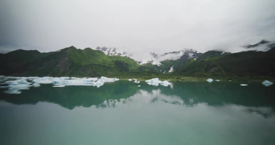 Glacial lake with floating ice blocks before green mountains with snow and low clouds reflected on the water