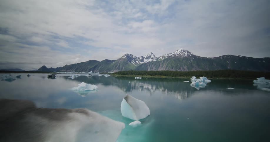 Ice block floating in a calm lake with snowcapped mountains in the background