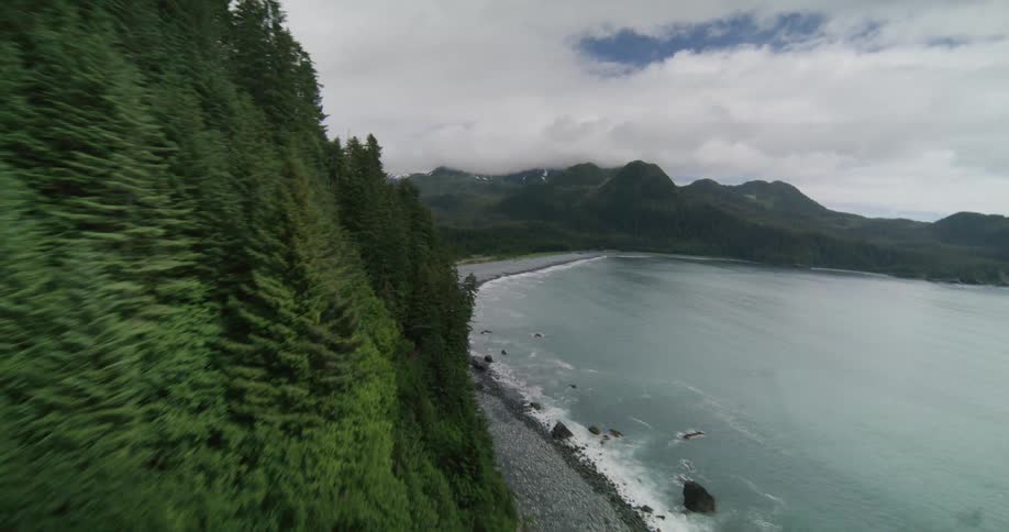 Aerial drone view flying along a steep green forested cliffside toward a secluded black sand beach and turquoise ocean bay under a cloudy sky