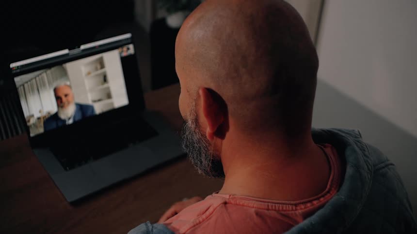 Man sitting at a table having a conversation with another man on a laptop computer