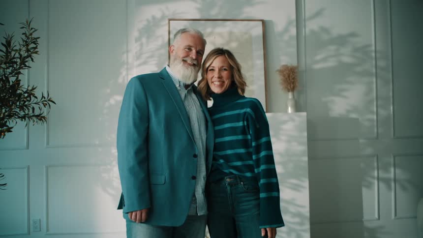 A bearded older man in a blue blazer and a woman in a striped turtleneck sweater stand smiling together in an interior space with wainscoting and natural light casting plant shadows on the wall