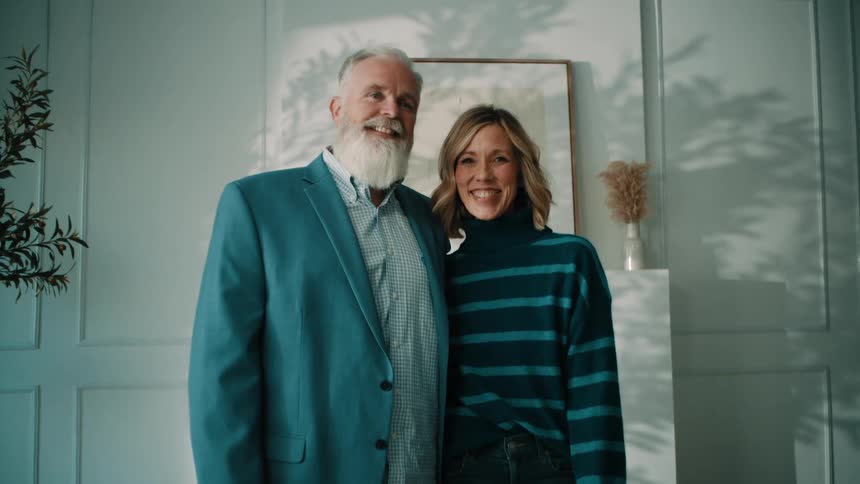 A smiling older man with a white beard and a blue blazer stands next to a woman in a striped turtleneck they are posing together in a brightly lit room with white paneled walls and a minimalist fireplace
