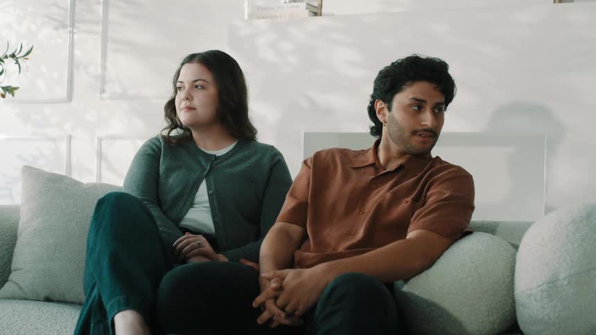 A couple sits side by side on a couch in their living room at midday sunlight filling the space around them