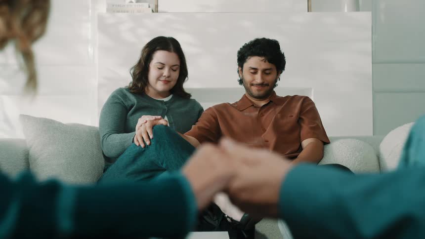 A closeup view shows two people holding hands while a young couple sits together on a couch in the background