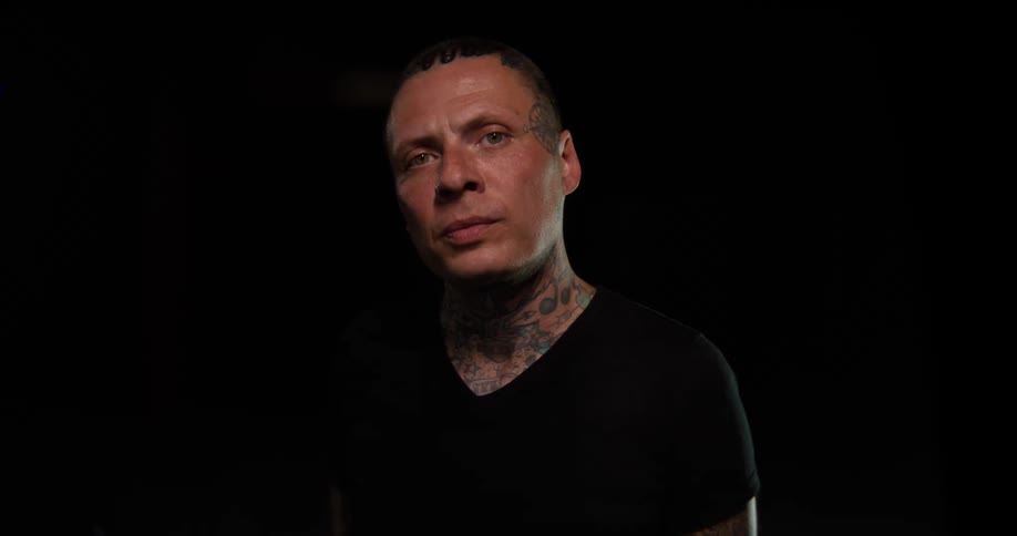 A man with numerous tattoos on his face and neck wearing a black t shirt looks directly ahead with a serious expression against a dark background