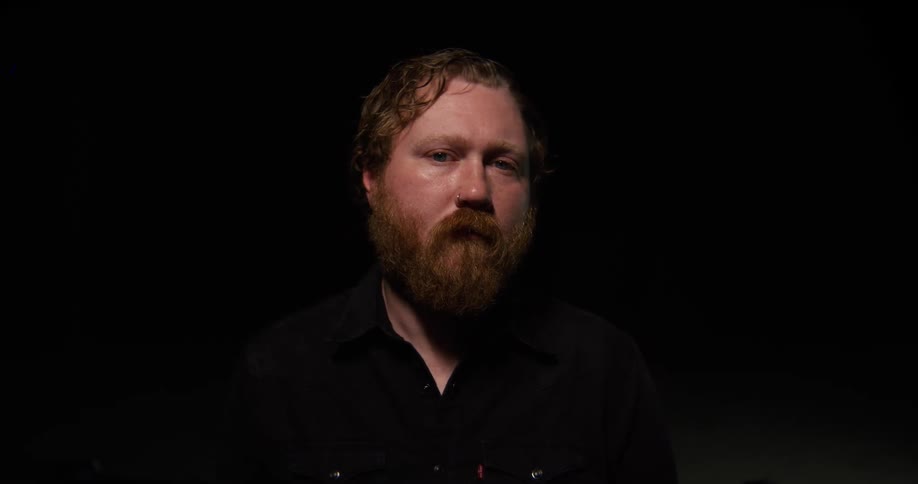Bearded man wearing a dark shirt faces forward under low lighting against a black background