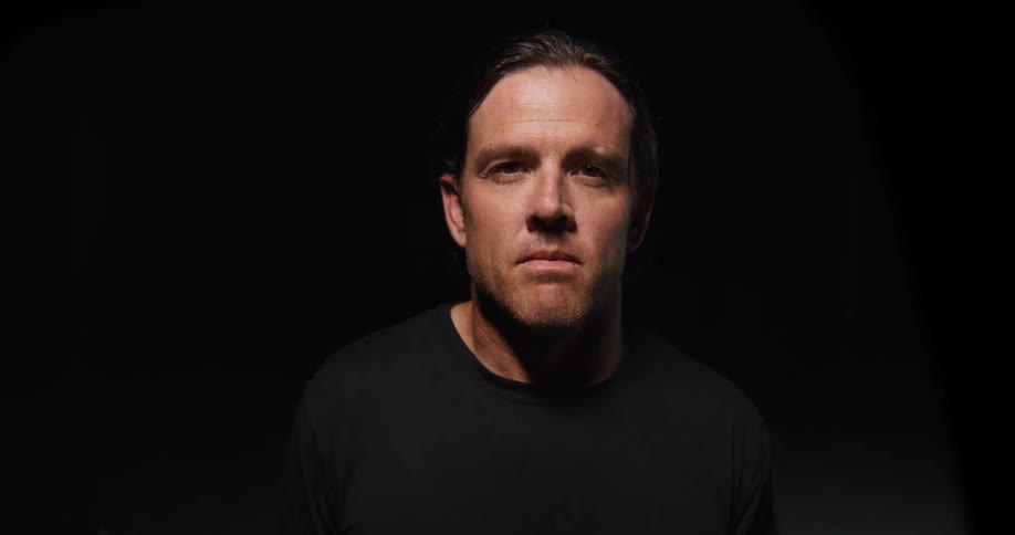 A man stands facing the camera in a dark studio under dramatic low key lighting