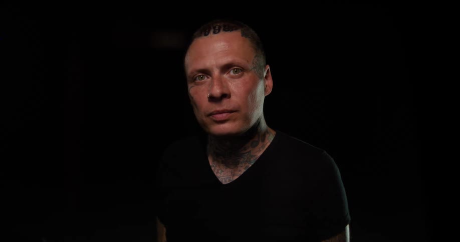 Man with neck tattoos wearing a black shirt faces forward under low lighting against a black background
