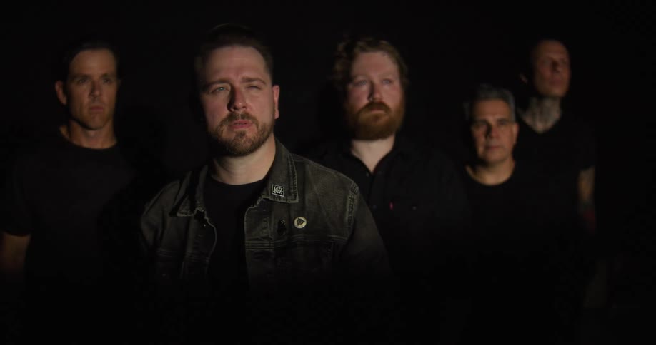 A group of five men stand together in a dark studio under dramatic lighting