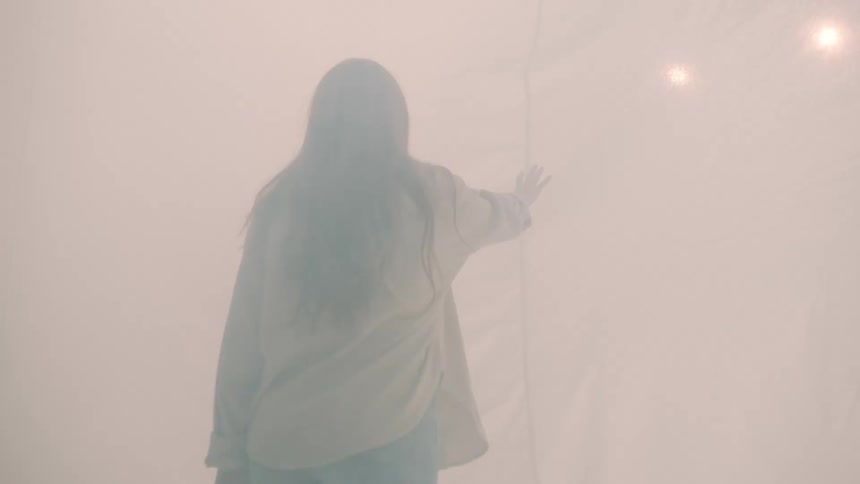 A young woman walks inside a tent pushing apart a linen divider as she tries to enter the next section bright flickering light shines through the fabric and the air around her is filled with drifting white smoke