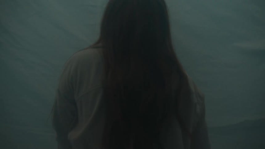 A young woman stands inside a room her long hair falling over her shoulders as the air around her fills with drifting indoor smoke