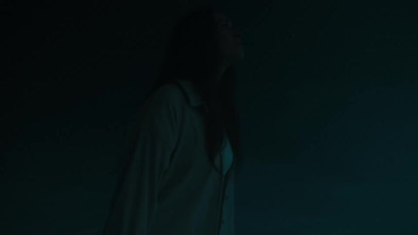 A young woman with long dark hair wears a white shirt and looks upward as blue light flashes against a dark background