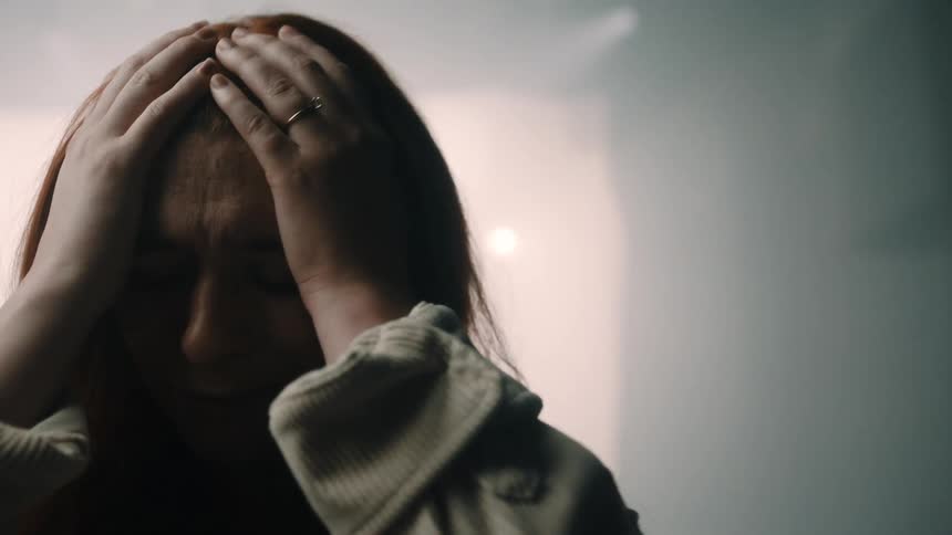 A close up shot shows a woman with long red hair as she closes her eyes and presses her hands against her head while standing in a dark space in front of a large white fabric sheet illuminated by bright lights