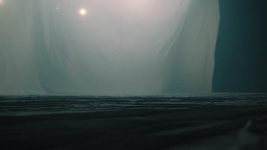 This video shows a lowangle shot of a dark misty room with large white fabric sheets draped in the background small warm glowing lights are visible through the haze creating a soft and atmospheric lighting effect