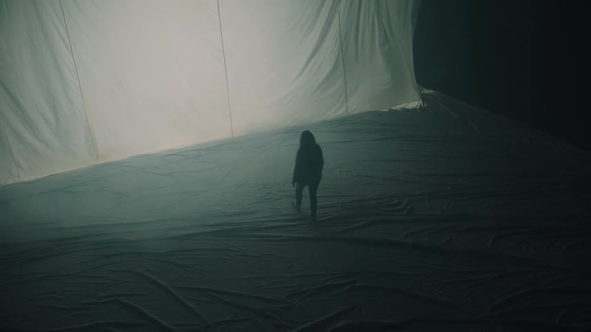 A high angle view shows a woman walking away from the camera toward a large white fabric sheet that is illuminated by bright lights in a dark space