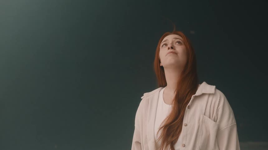 A woman with long red hair and a white jacket looks up the background is a dark blue wall with white particles falling in the air