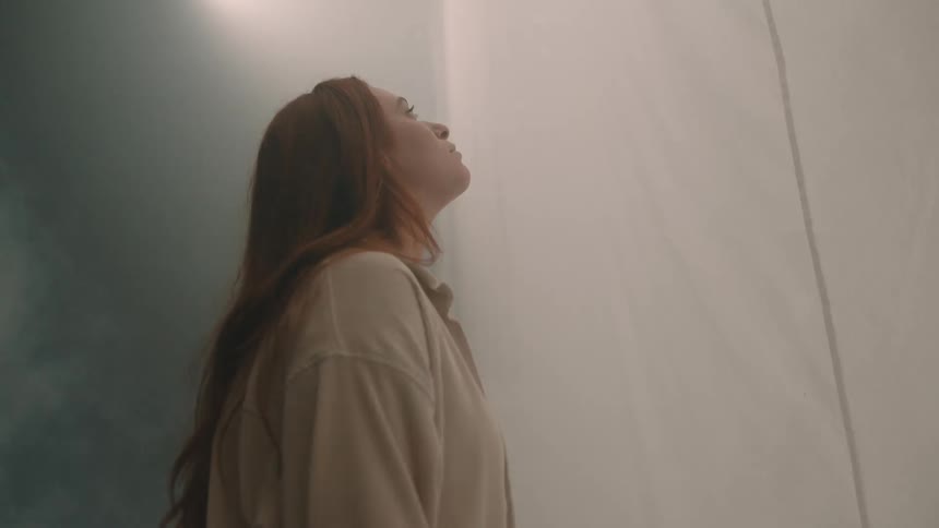 Young adult woman standing and staring at a white curtain under a spolight surrounded by smoke indoors