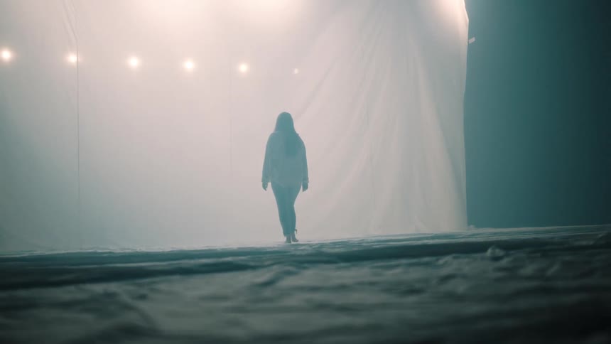 A young woman in a white shirt and jeans walks away from the camera toward a large white fabric sheet illuminated by bright lights in a dark space