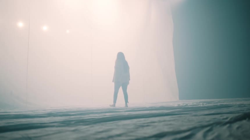 A young woman with long red hair walks through a large tent filled with white smoke moving toward a bright light ahead